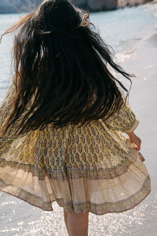 Back view of woman with windswept hair on a beach wearing the Daughters of India Bhoomi Mini Dress in Meadow, showing the tiered skirt and beautiful border hem blowing in the breeze