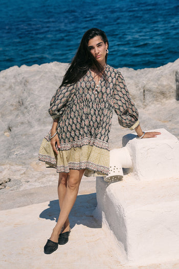 Woman standing on white stone by the ocean wearing the Daughters of India Bhoomi Mini Dress in Onyx, a dark block print cotton mini dress with tiered skirt and billowed sleeves