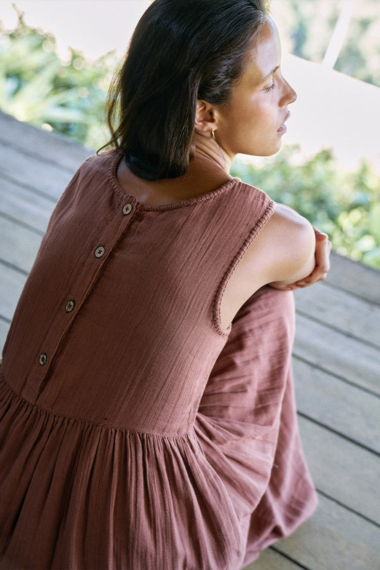 Nadi Dress Chocolate — close-up back view seated on wooden deck, back button detail and gathered waist, chocolate brown double gauze cotton, tropical greenery background | Daughters of India