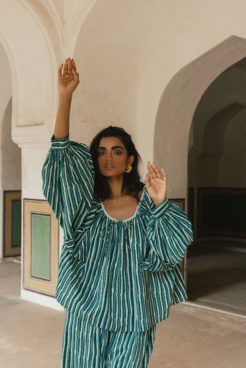 Ria Blouse Cyan Candy — back view in architectural setting, model showing rear of cyan and dark stripe hand block print cotton blouse, relaxed fit with peplum hem detail, artisan Jaipur India | Daughters of India