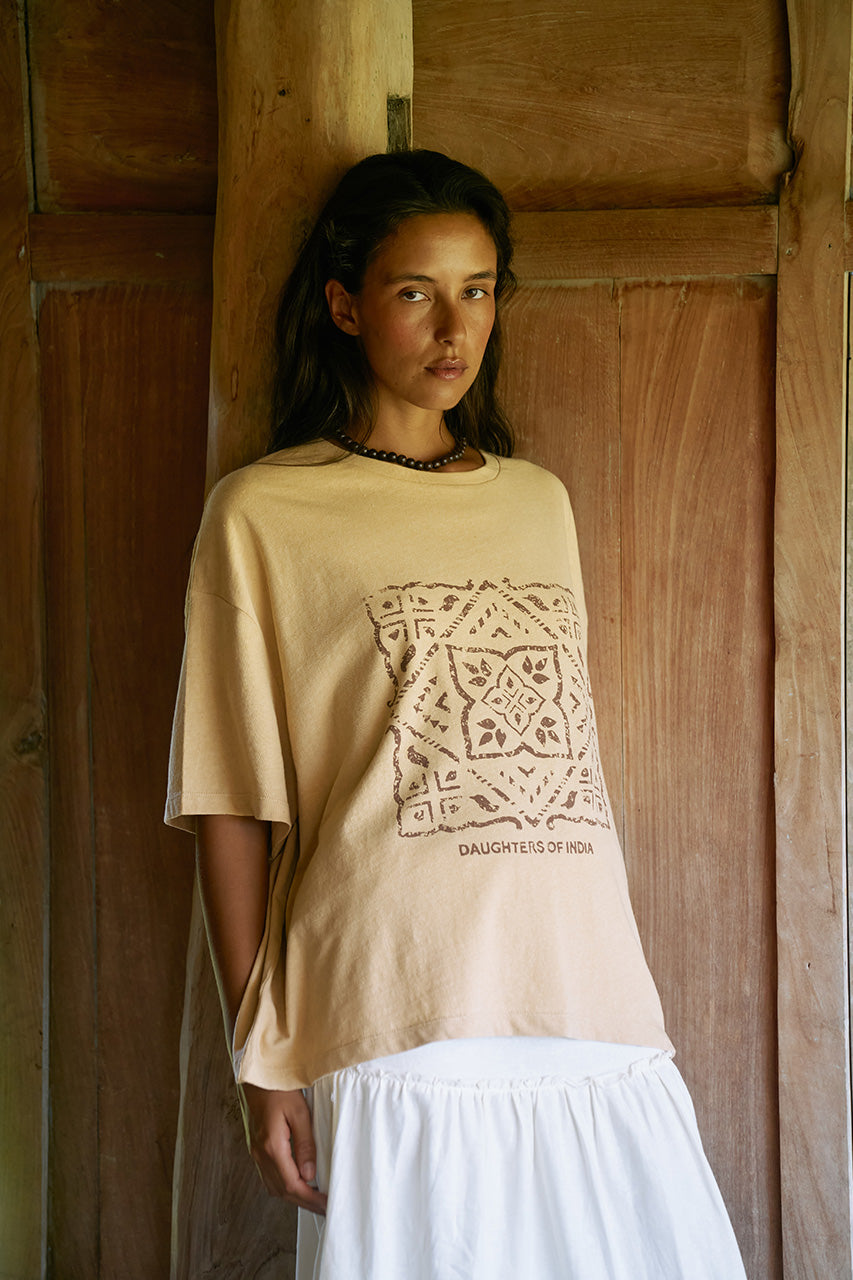 Woman standing against a wooden door wearing the Daughters of India Shanti Tee in Cashew with a white ruffled skirt and beaded necklace