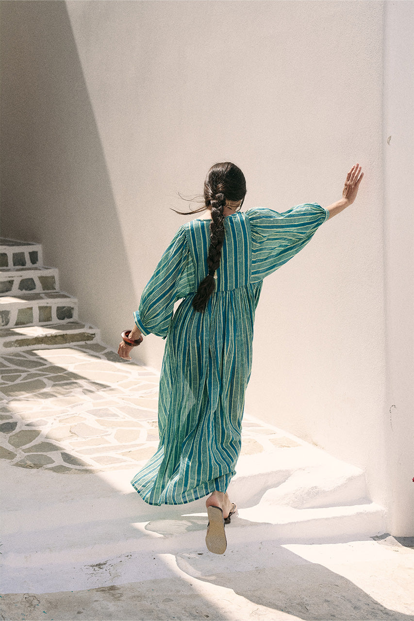 Back view of woman walking up white steps wearing the Daughters of India Kyra Maxi Dress in Spearmint, showing the flowing back of the green striped dress with braided hair