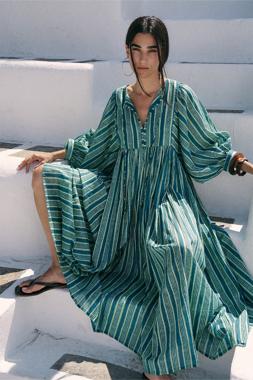 Woman seated on white steps wearing the Daughters of India Kyra Maxi Dress in Spearmint, showing the voluminous billowed sleeves and flowing green striped fabric